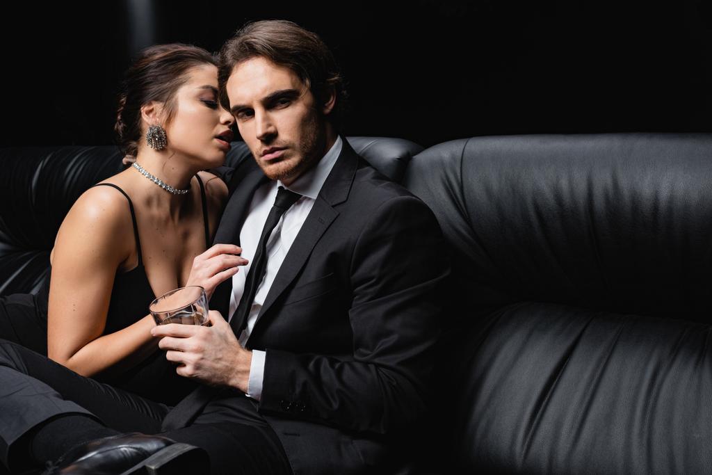 sexy Frau im Slip-Kleid verführt Mann im Anzug hält Glas Whiskey auf schwarz  - Foto, Bild