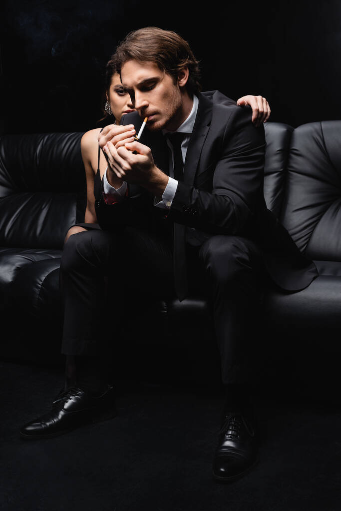 жінка в сукні ковзання, що обіймає чоловіка в костюмі, тримає запальничку біля цигарки на чорному
 - Фото, зображення