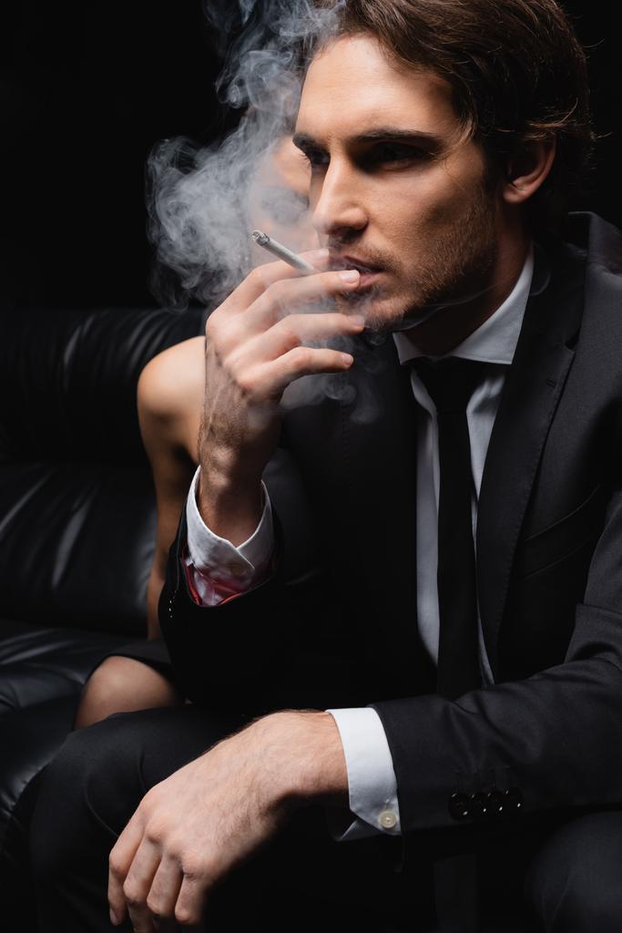 serious man in suit smoking cigarette near woman on blurred black background  - Photo, Image