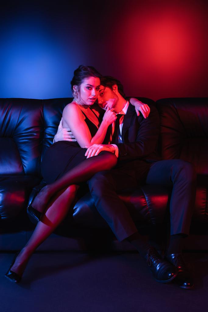 éclairage rouge et bleu sur sexy homme et femme étreignant sur canapé en cuir noir  - Photo, image