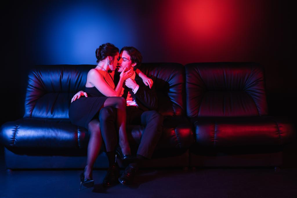 pełna długość czerwono-niebieskie oświetlenie na sexy mężczyzna i kobieta całuje na czarnej skórzanej kanapie  - Zdjęcie, obraz