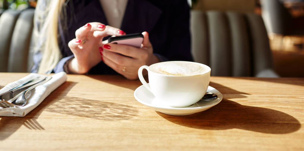 Kobieta pisząca SMS-y na smartfonie w kawiarni. Młoda kobieta siedząca przy stole z kawą przy użyciu telefonu komórkowego. Przerwa na kawę. Filiżanka kawy na stole. Niewyraźny obraz, selektywne skupienie - Zdjęcie, obraz