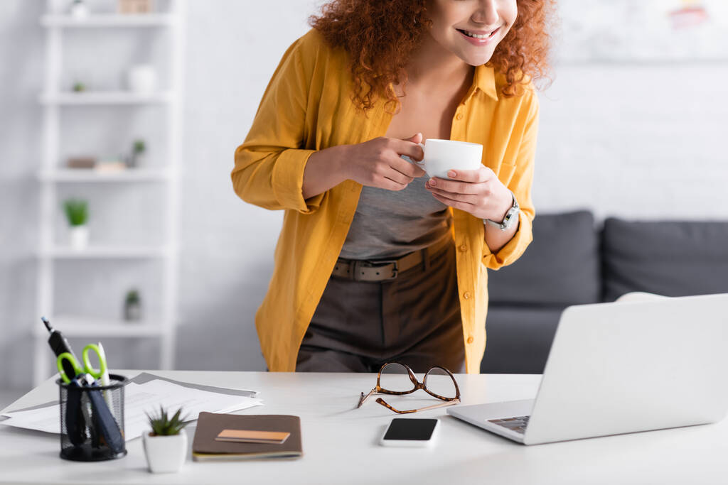обрезанный вид фрилансера, держащего чашку кофе, стоя рядом с ноутбуком - Фото, изображение