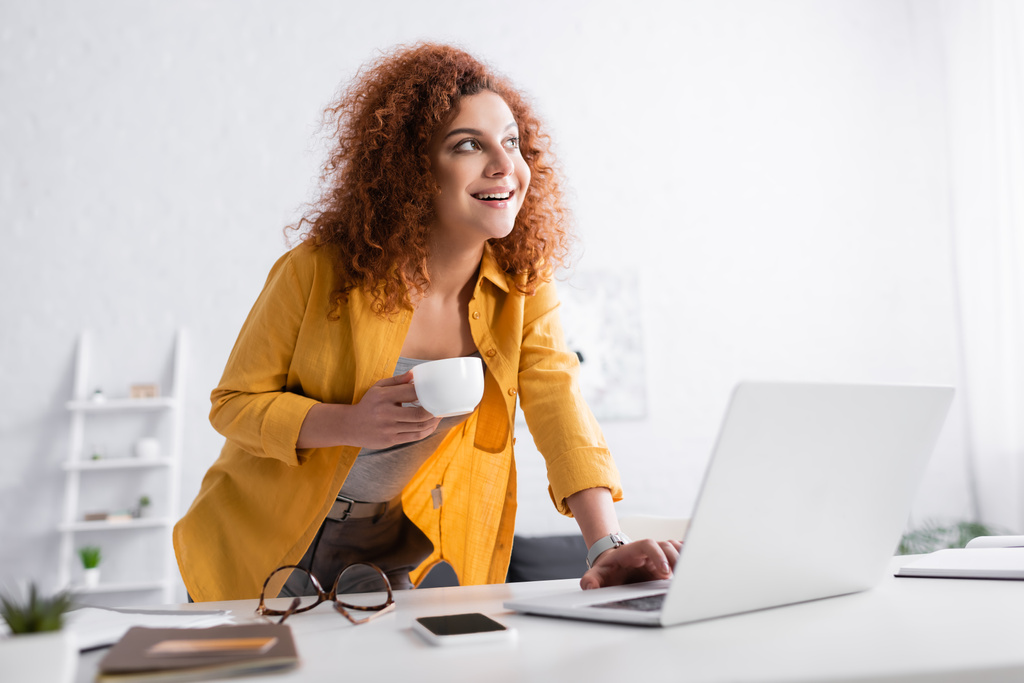 alegre freelancer de pie con taza de café cerca de la computadora portátil en primer plano borrosa - Foto, Imagen