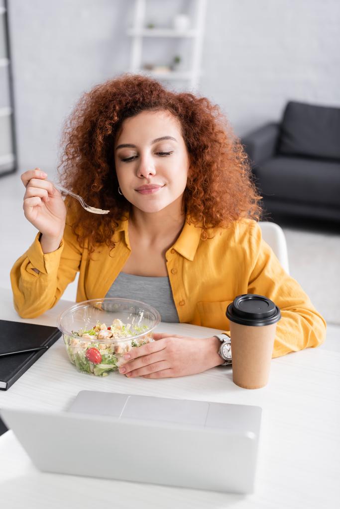 curly freelancer eating fresh salad near laptop on blurred foreground - Photo, Image