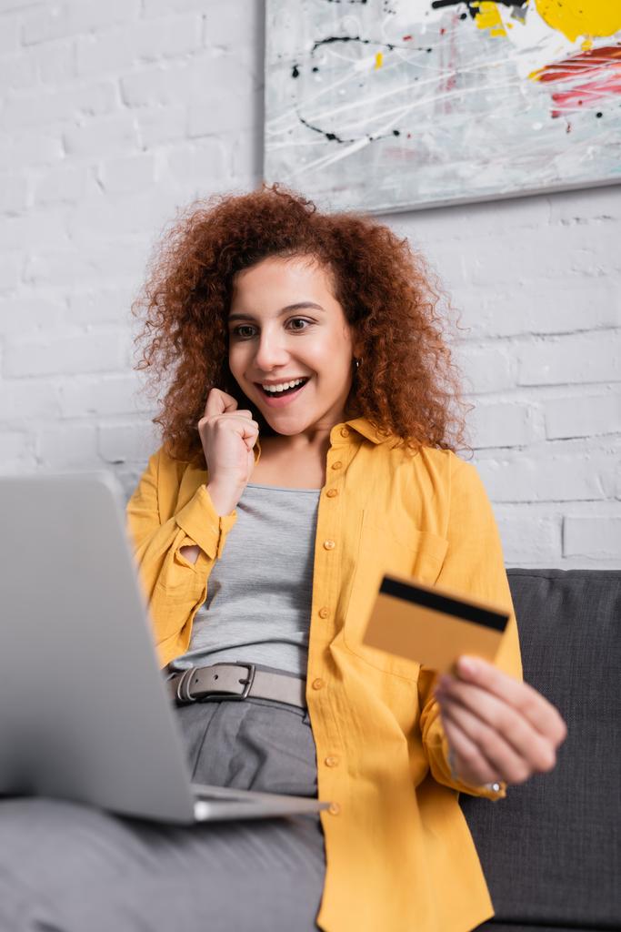счастливая женщина держит кредитную карту возле ноутбука на размытом переднем плане - Фото, изображение