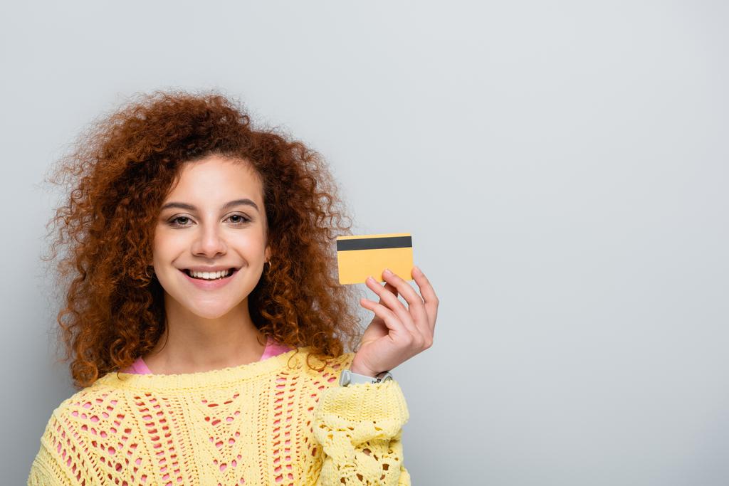 Радостная женщина в свитере с открытыми дверями, держащая кредитную карту изолированную на сером - Фото, изображение