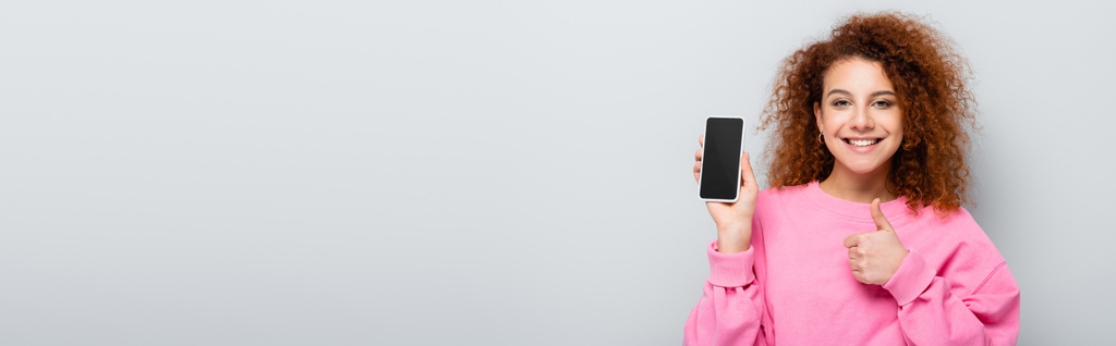 fröhliche Frau zeigt Daumen hoch, während sie Handy mit leerem Bildschirm auf grau isoliert hält, Banner - Foto, Bild
