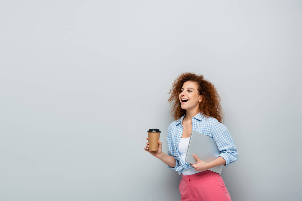 amazing woman carrying coffee to go and laptop on grey background - Photo, Image