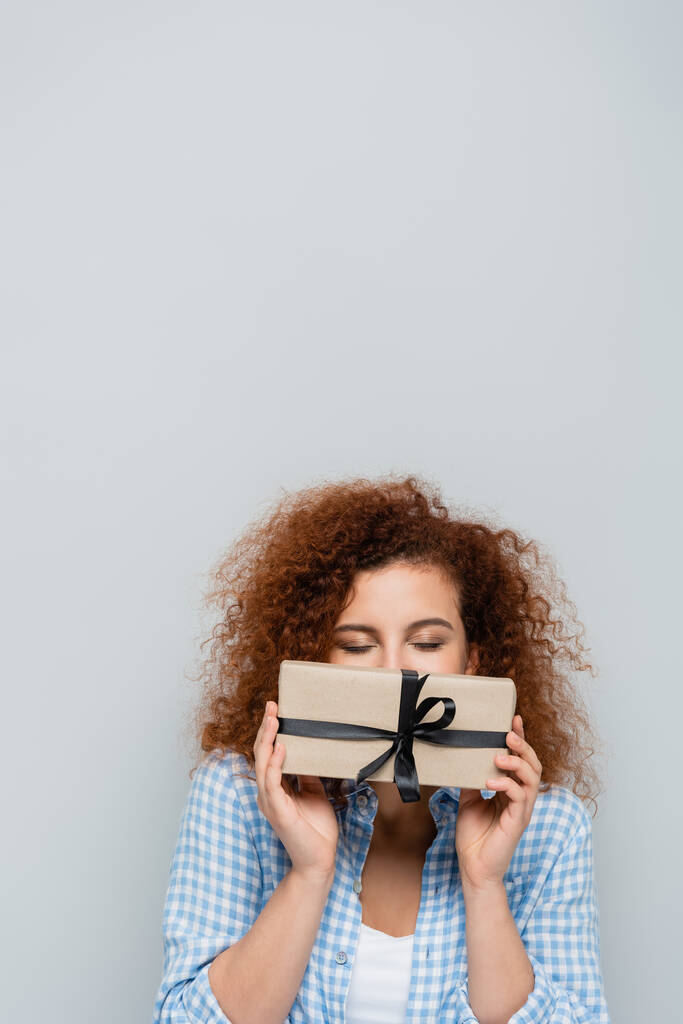 кудрявая женщина с закрытыми глазами затемняющее лицо с подарочной коробкой, изолированной на сером - Фото, изображение