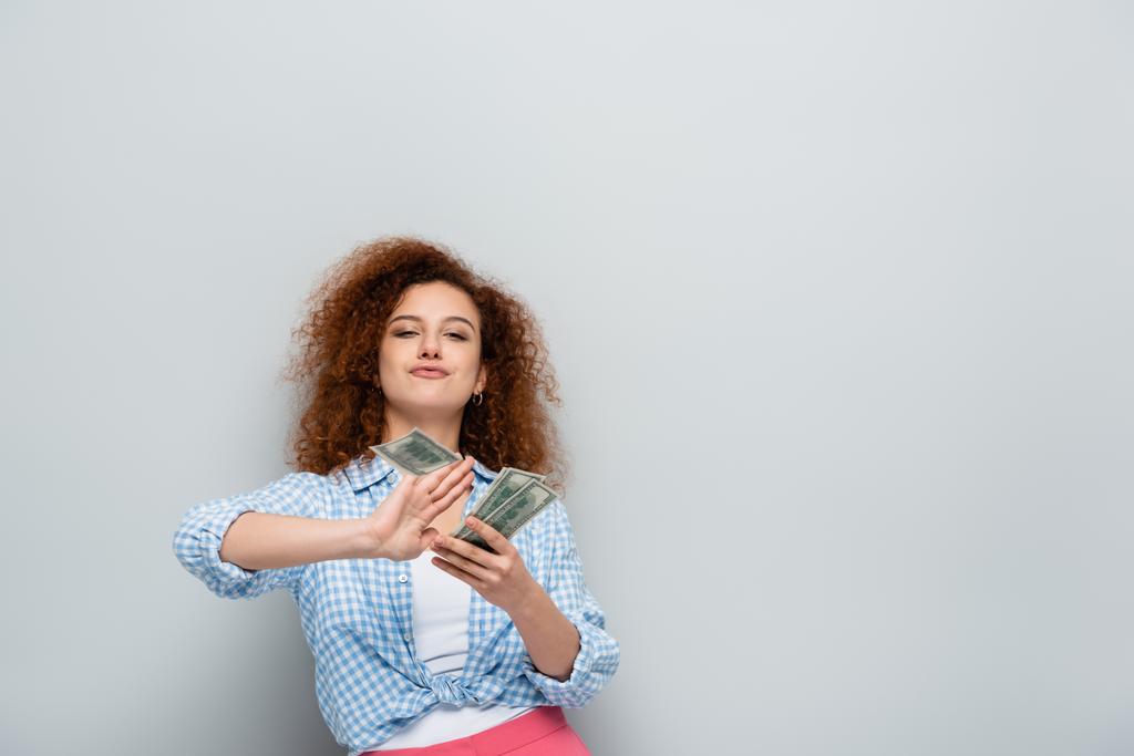mulher alegre contando dólares enquanto olha para a câmera no fundo cinza - Foto, Imagem