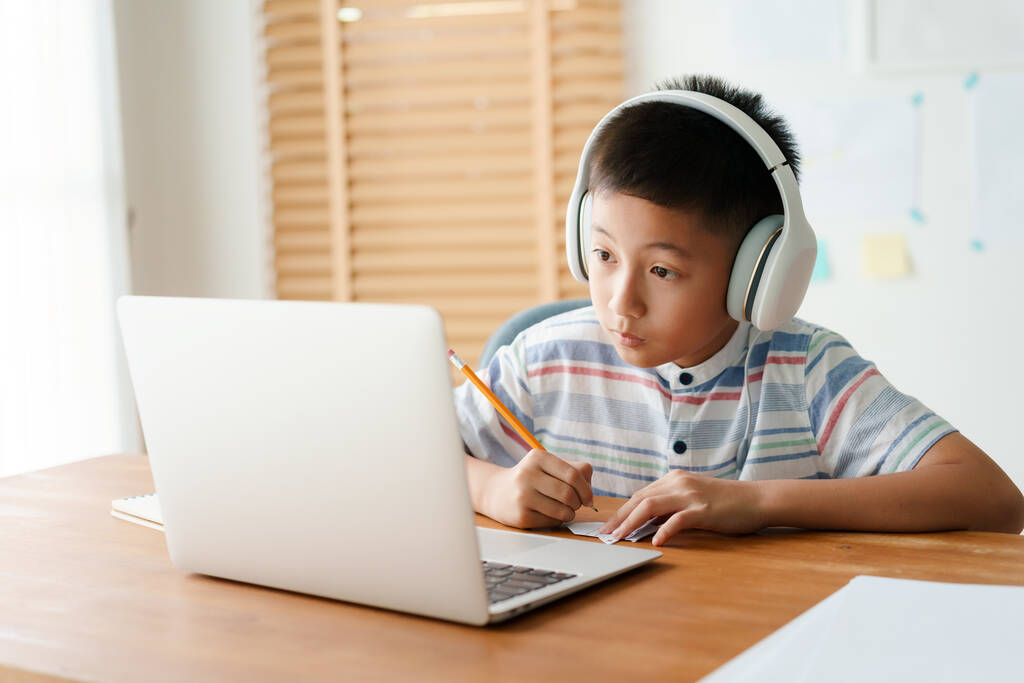 Asyalı küçük bir çocuk internetten bilgisayar hocasıyla kulaklıklı bir bilgisayar laptopuyla video görüşmesi yapıyor. Asyalı ev ödevi yapıyor. Evde online öğrenme kavramını kabul et  - Fotoğraf, Görsel