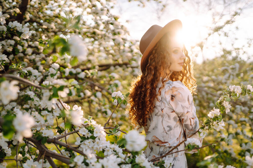 Mulher encaracolada com o chapéu posando no parque de primavera florescendo. O conceito de relaxar, viajar, paraíso, romance e férias de primavera. Estilo de moda. - Foto, Imagem