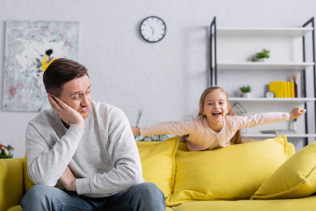 Отец смотрит на позитивного ребенка рядом с диваном дома  - Фото, изображение