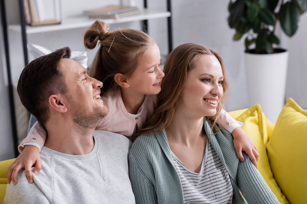 Lächelndes Mädchen umarmt Eltern auf gelber Couch  - Foto, Bild