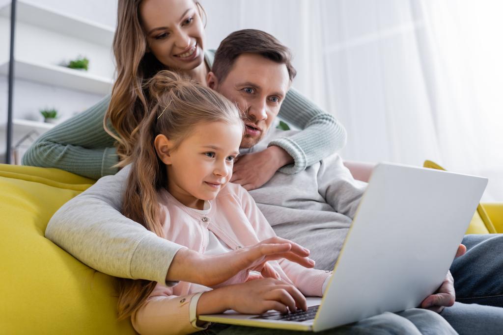 Homme utilisant un ordinateur portable avec sa fille près d'une femme souriante sur fond flou  - Photo, image