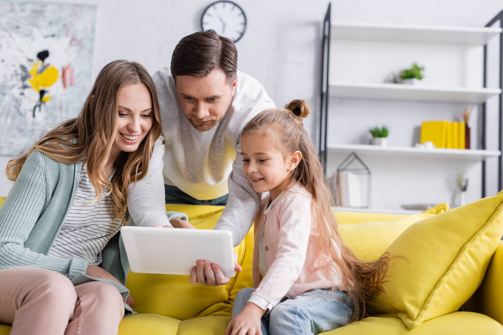 Mann benutzt digitales Tablet neben lächelnder Frau und Tochter auf Couch  - Foto, Bild