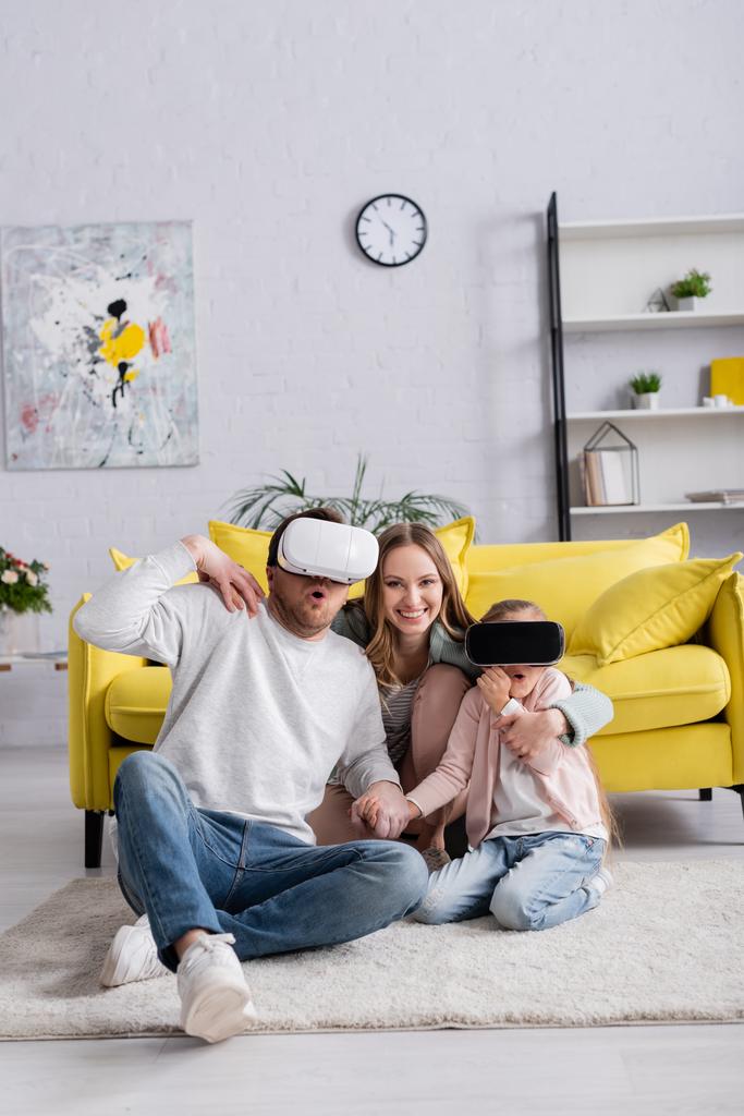Lächelnde Frau sitzt mit Kopfhörern neben Mann und Tochter auf Teppich  - Foto, Bild