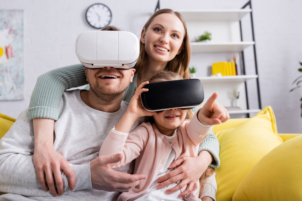 Uśmiechnięta kobieta przytula córkę i męża w słuchawkach VR na kanapie  - Zdjęcie, obraz