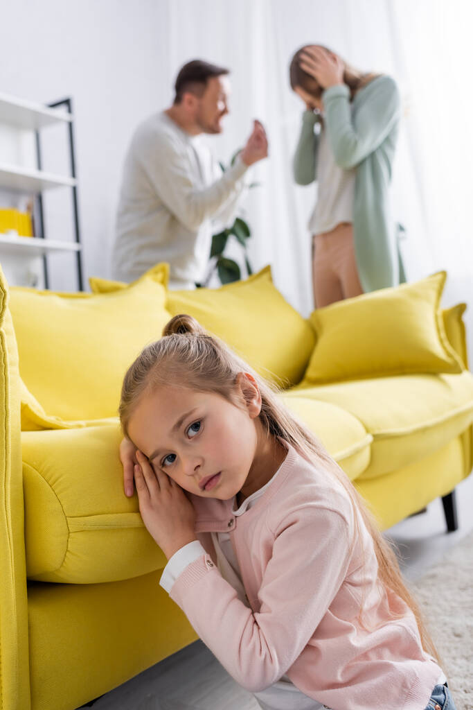 Dívka při pohledu na kameru v blízkosti gauče a rodiče mají konflikt na rozmazaném pozadí  - Fotografie, Obrázek