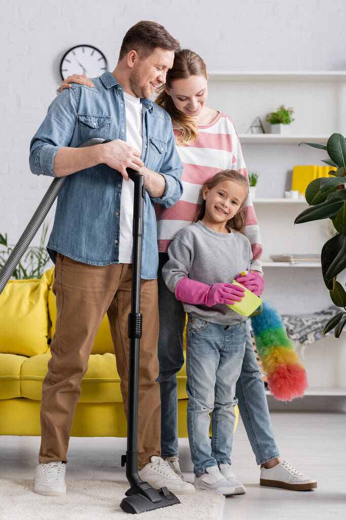 Famiglia sorridente con prodotti per la pulizia in piedi in soggiorno  - Foto, immagini