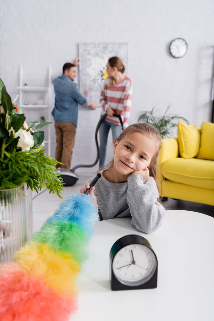 Sonriente niño con cepillo de polvo mirando a la cámara cerca del reloj y las plantas en la mesa  - Foto, Imagen