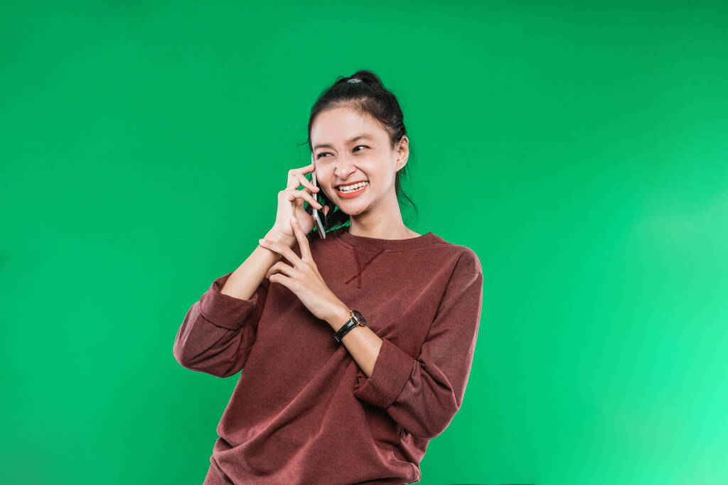 hermosa joven asiática mujer está hablando por teléfono sonriendo débilmente con alguien mientras mira al lado - Foto, Imagen