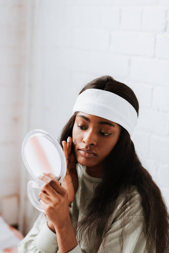 Una atractiva joven africana con pelo largo aplicando crema facial mientras se mira en un espejo de mano en el dormitorio - Foto, imagen