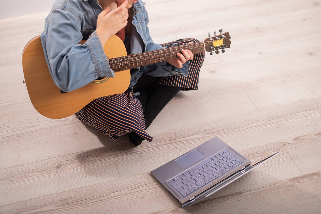 Młoda hipsterka siedząca w domu na podłodze i oglądająca poradniki gitarowe na laptopie. Muzyka do nauki na odległość w kwarantannie - Zdjęcie, obraz