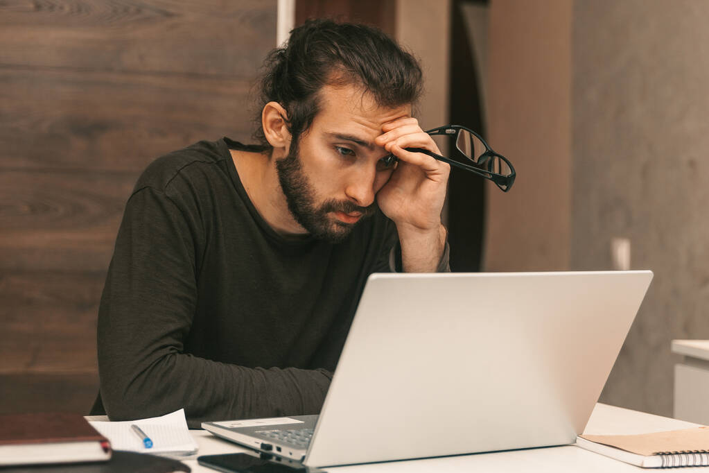 Mladík se unaveně podívá na laptop. Práce z domova. Muž pracující na notebooku - Fotografie, Obrázek