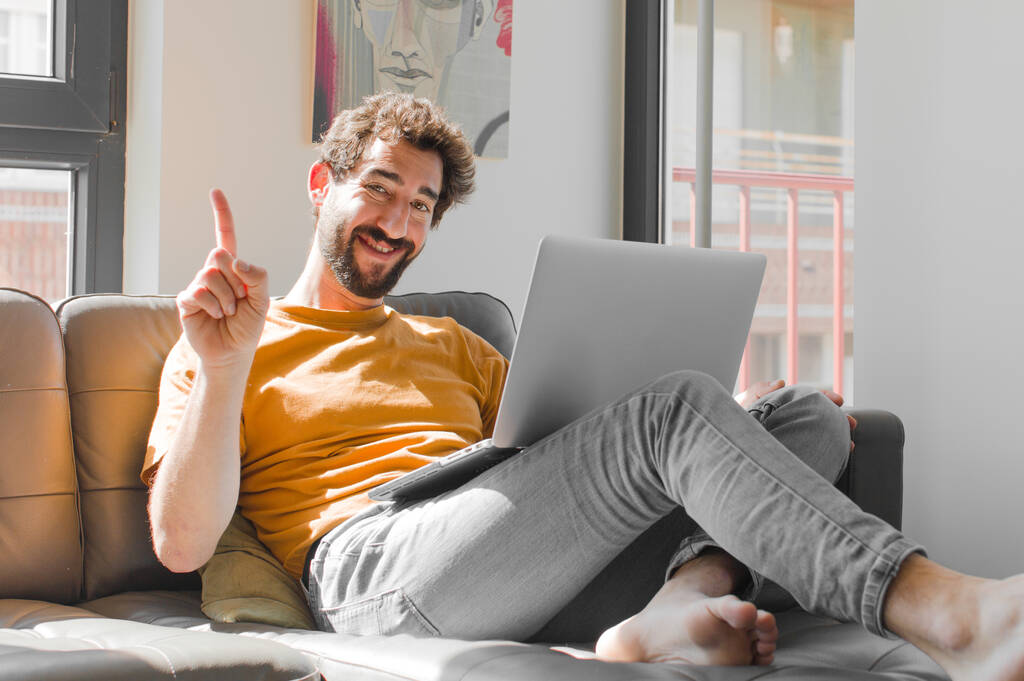 молодий бородатий чоловік посміхається і виглядає доброзичливо, показуючи номер один або спочатку з рукою вперед, рахуючи з ноутбуком на дивані концепції ноутбука
 - Фото, зображення