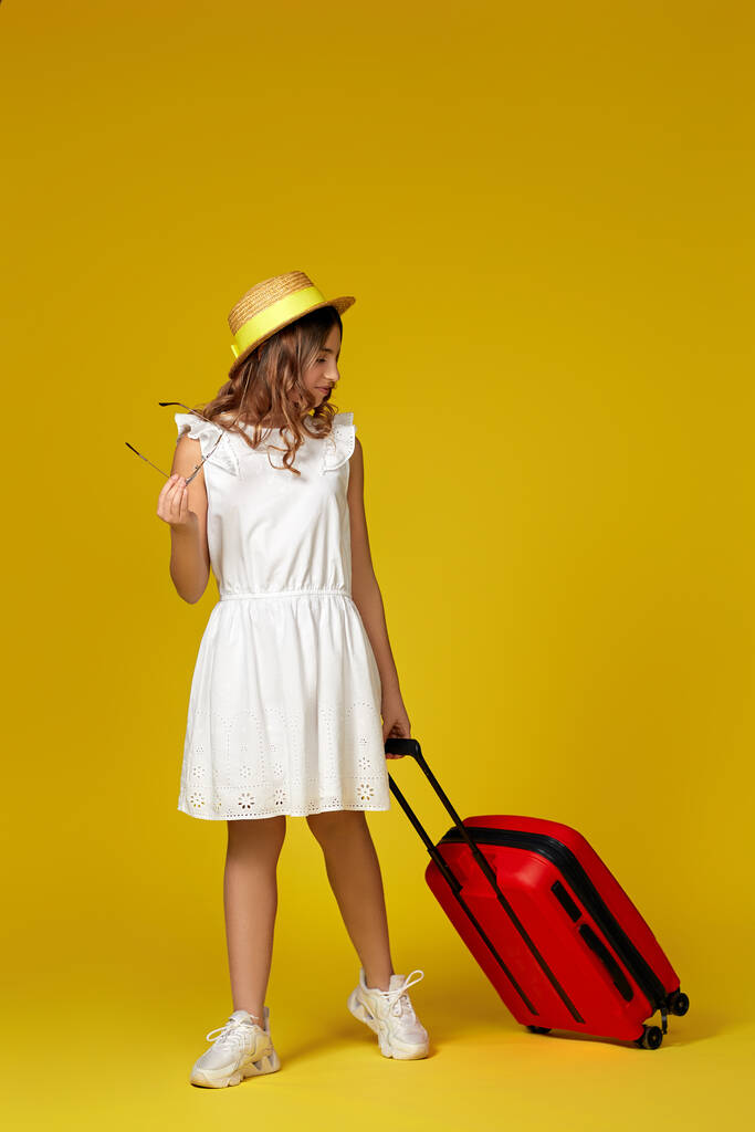 девушка-подросток в шляпе и солнечных очках с красным чемоданом - Фото, изображение