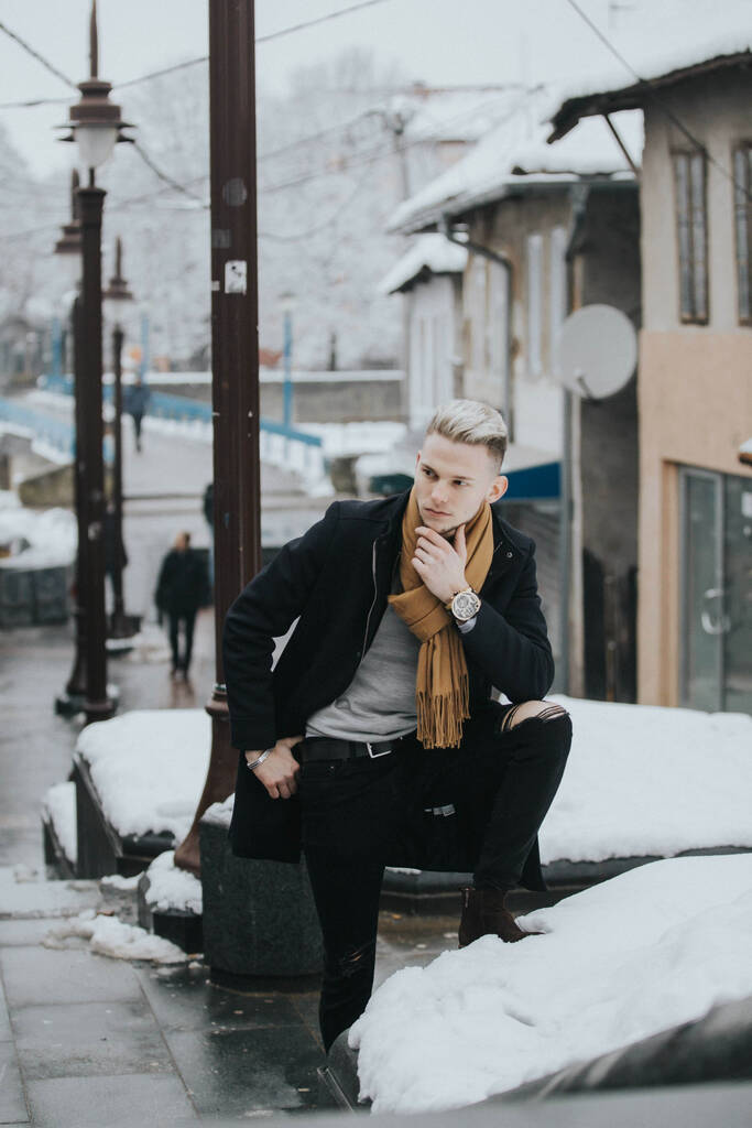 Egy jóképű, magabiztos férfi fekete téli ruhában, barna sállal a lábával a támfalon. - Fotó, kép
