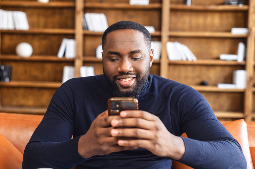 Αφροαμερικάνος κάθεται με το τηλέφωνο στα χέρια παθιασμένος με κάτι - Φωτογραφία, εικόνα