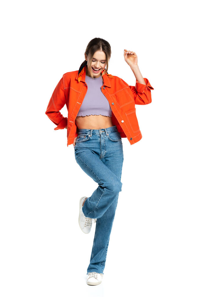 volledige lengte van positieve jonge vrouw in gewas top, jeans en oranje shirt poseren geïsoleerd op wit  - Foto, afbeelding