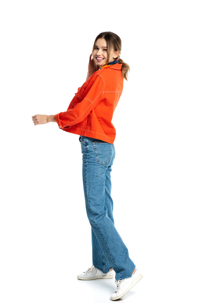 longitud completa de la mujer joven positiva en jeans y camisa naranja de pie aislado en blanco  - Foto, Imagen