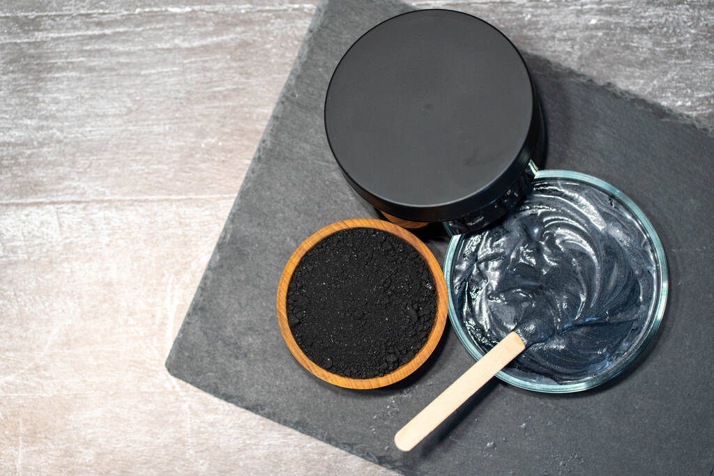 Домашняя маска для лица и скраб с активированным угольным порошком и йогуртом с палкой на черной тарелке с баночкой. - Фото, изображение