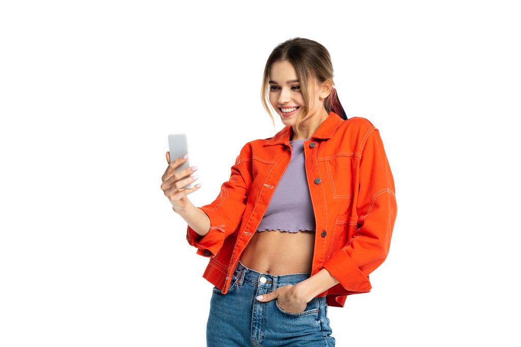 gelukkig jong vrouw in jeans en oranje shirt het nemen van selfie terwijl staan met de hand in zak geïsoleerd op wit - Foto, afbeelding