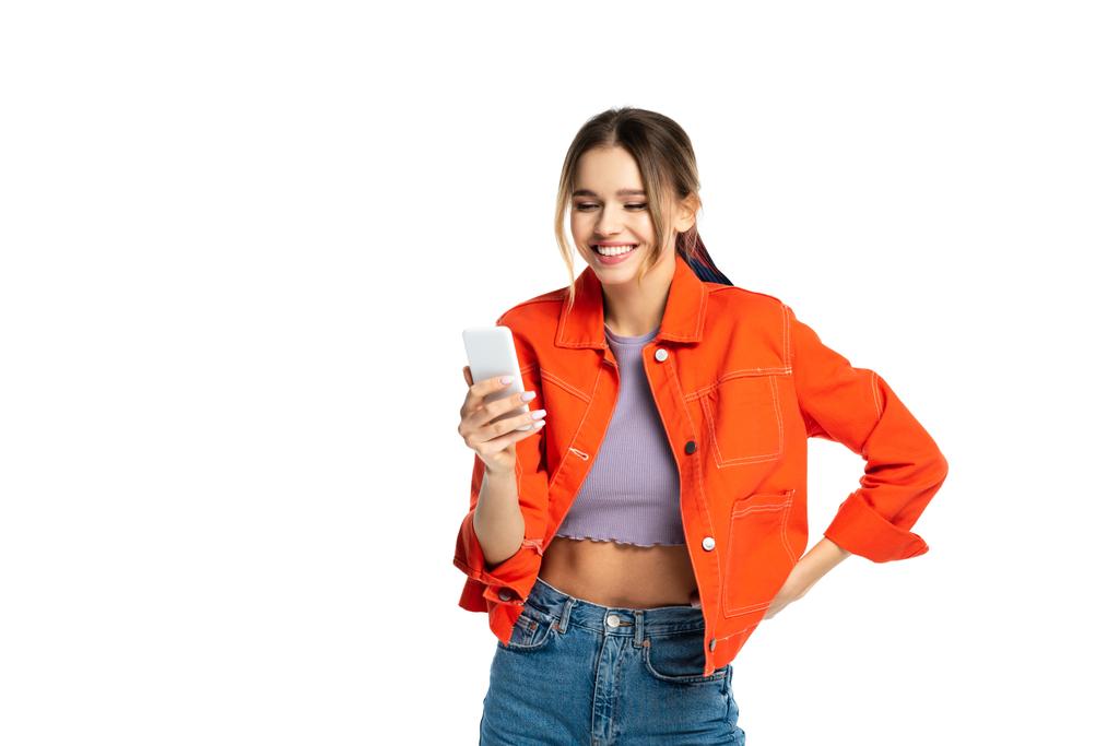 mujer joven feliz en la parte superior de la cosecha y camisa naranja usando el teléfono inteligente, mientras que de pie con la mano en la cadera aislado en blanco - Foto, Imagen