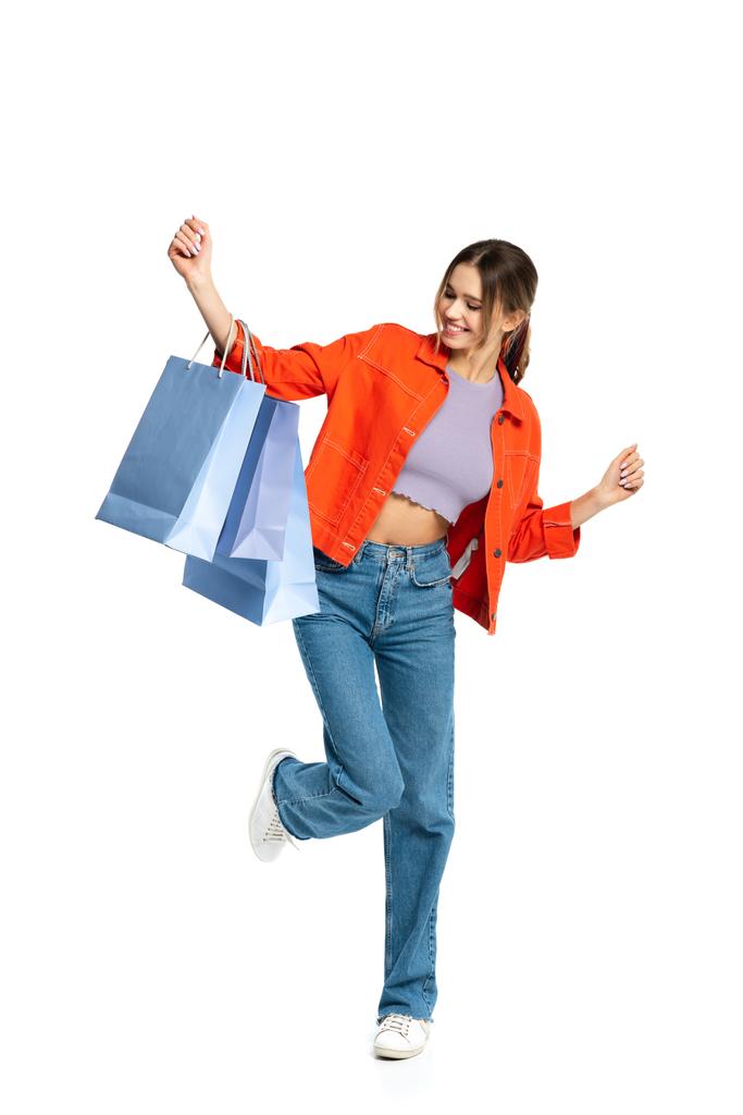 オレンジ色のシャツを着た幸せな若い女性の完全な長さは白で隔離されたショッピングバッグを見て  - 写真・画像