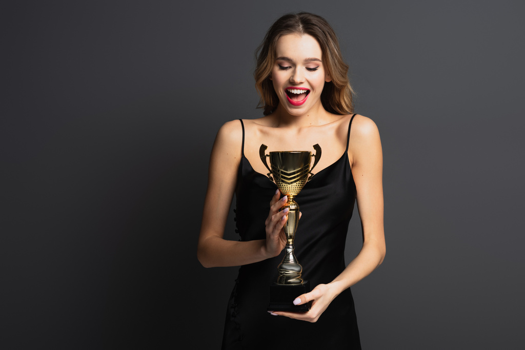 excitée jeune femme en robe noire glissante tenant trophée d'or sur gris - Photo, image