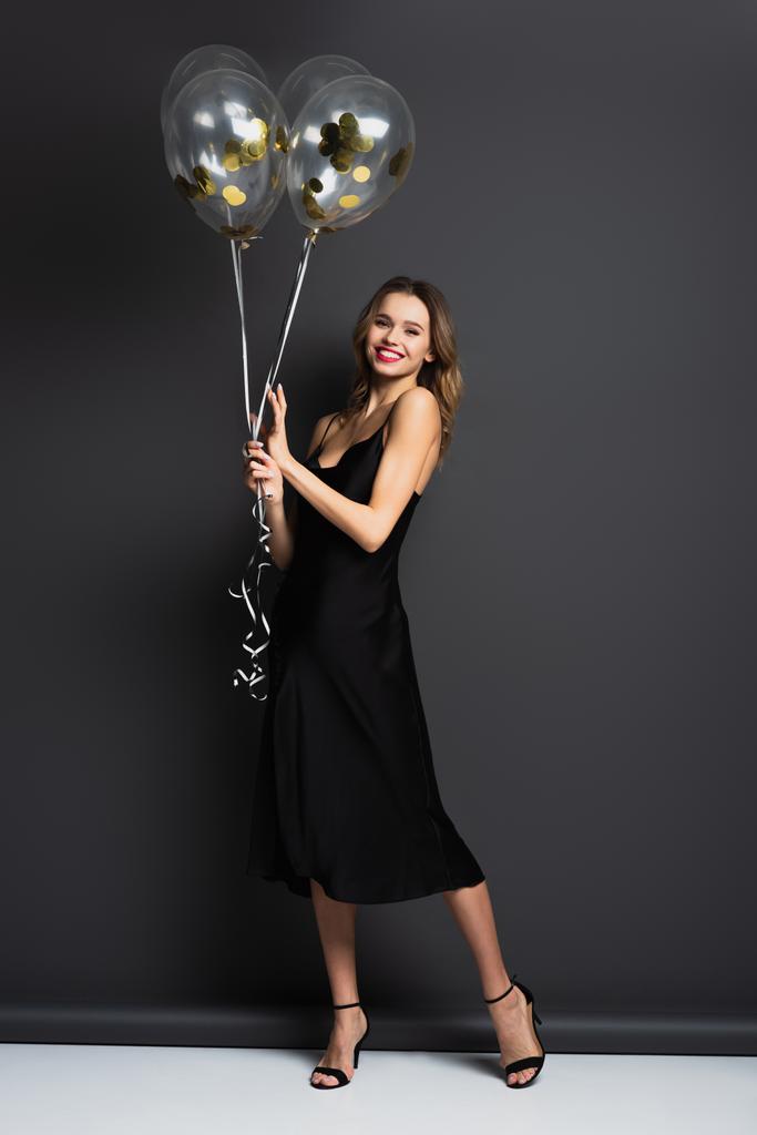 volledige lengte van glimlachende jonge vrouw in zwarte slip jurk houden ballonnen op grijs - Foto, afbeelding