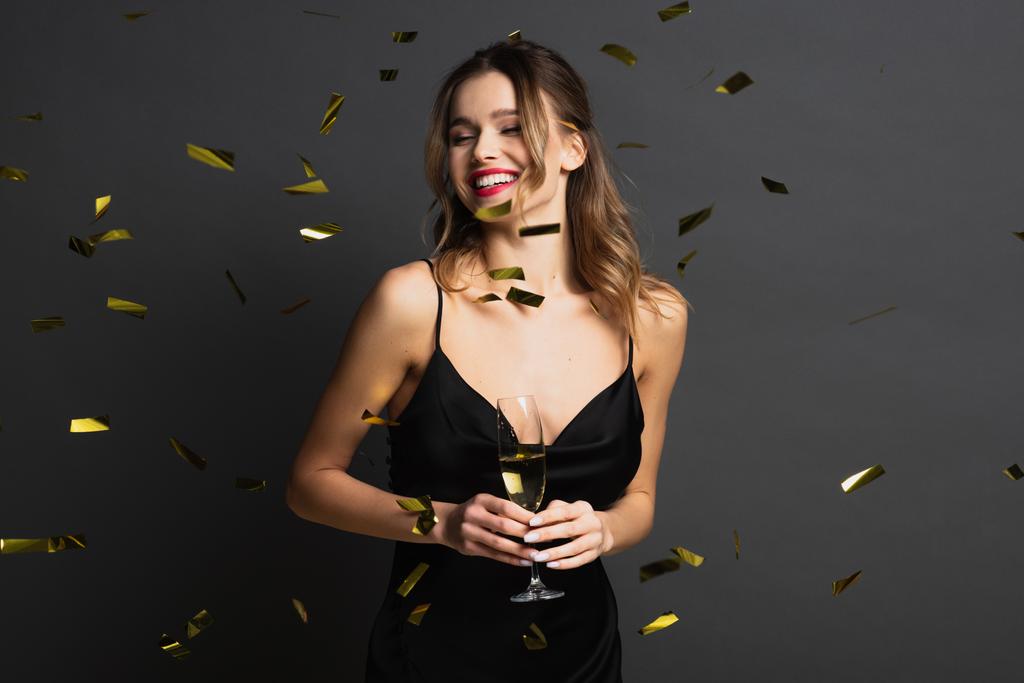 vreugdevolle jonge vrouw in zwarte slip jurk met glas champagne in de buurt confetti op grijs - Foto, afbeelding