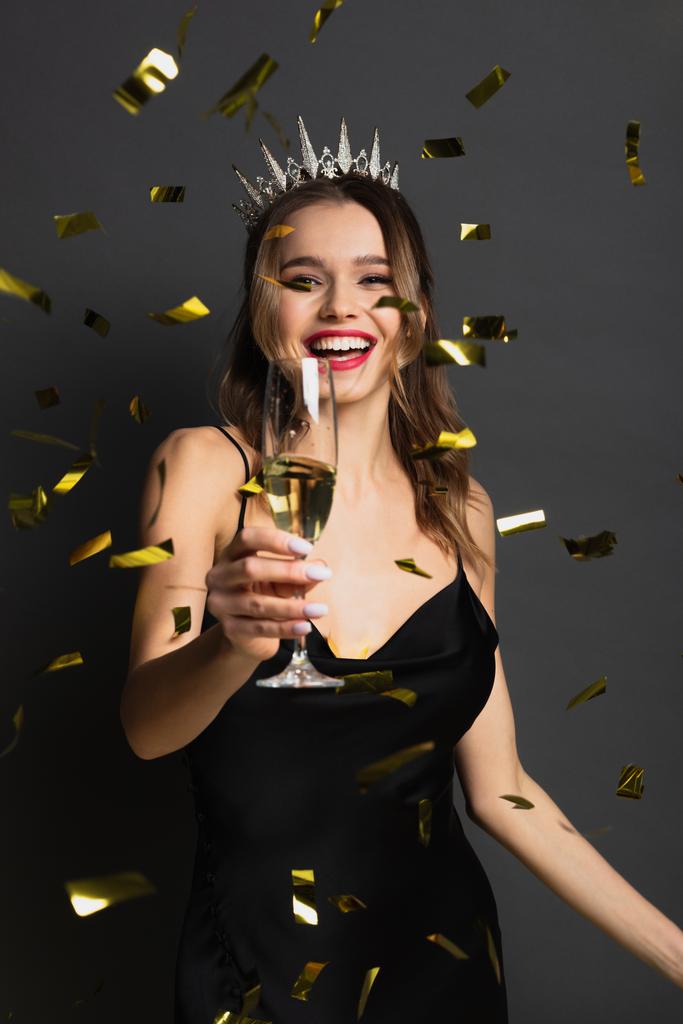 radosna kobieta w poślizgowej sukience i tiara trzymająca kieliszek szampana w pobliżu confetti na szarości - Zdjęcie, obraz