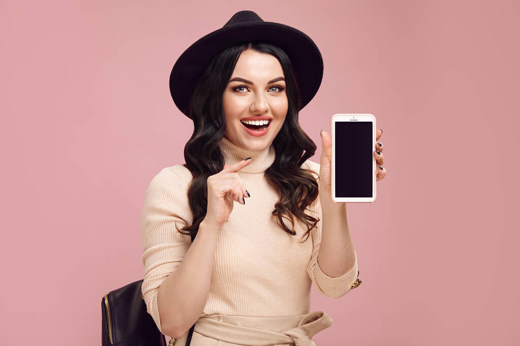 Chica adulta alegre señala a la pantalla en blanco de su teléfono y sonríe sorprendido mientras está de pie sobre rosa aislado - Foto, imagen