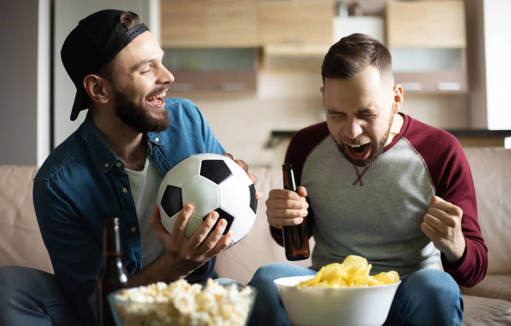 Αστεία ενθουσιασμένοι hipster γενειοφόροι φίλοι με σνακ και μπύρες βλέποντας ποδόσφαιρο στην τηλεόραση, ενώ κάθεται στον καναπέ στο σπίτι. Οι οπαδοί φωνάζουν λόγω της νίκης της ομάδας τους - Φωτογραφία, εικόνα