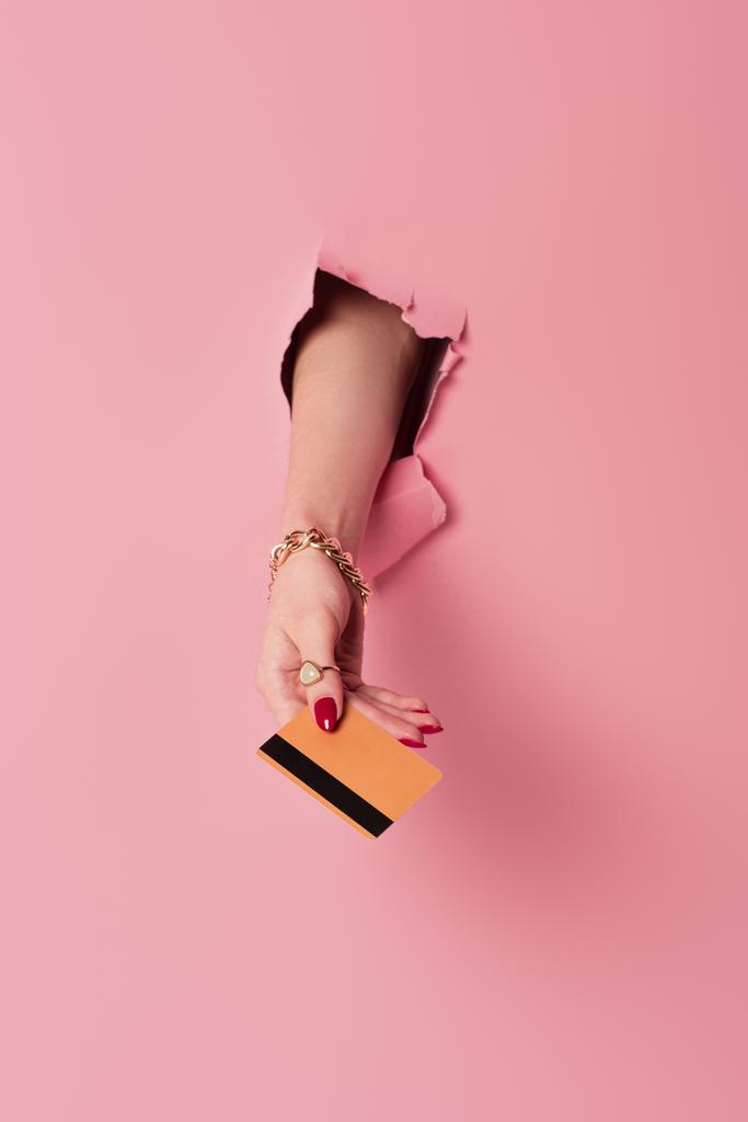 Cropped view of woman with bracelet and ring holding credit card in hole on pink background - Photo, Image