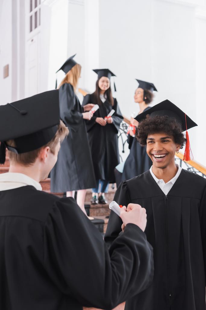 Glimlachende Afrikaans-Amerikaanse afgestudeerde met diploma staan in de buurt vriend op wazig voorgrond  - Foto, afbeelding