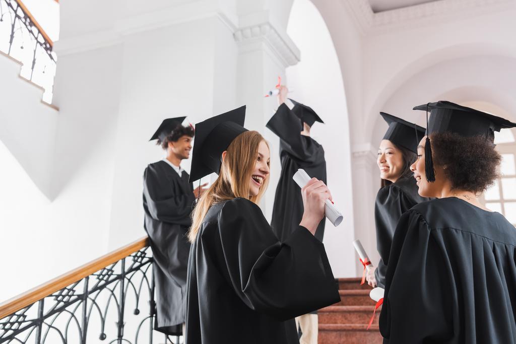 Lachende afgestudeerde in cap holding diploma in de buurt van interraciale vrienden in de universiteit  - Foto, afbeelding