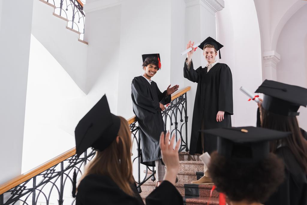 Graduados multiétnicos sonrientes con diplomas saludando a sus amigos en un primer plano borroso  - Foto, Imagen